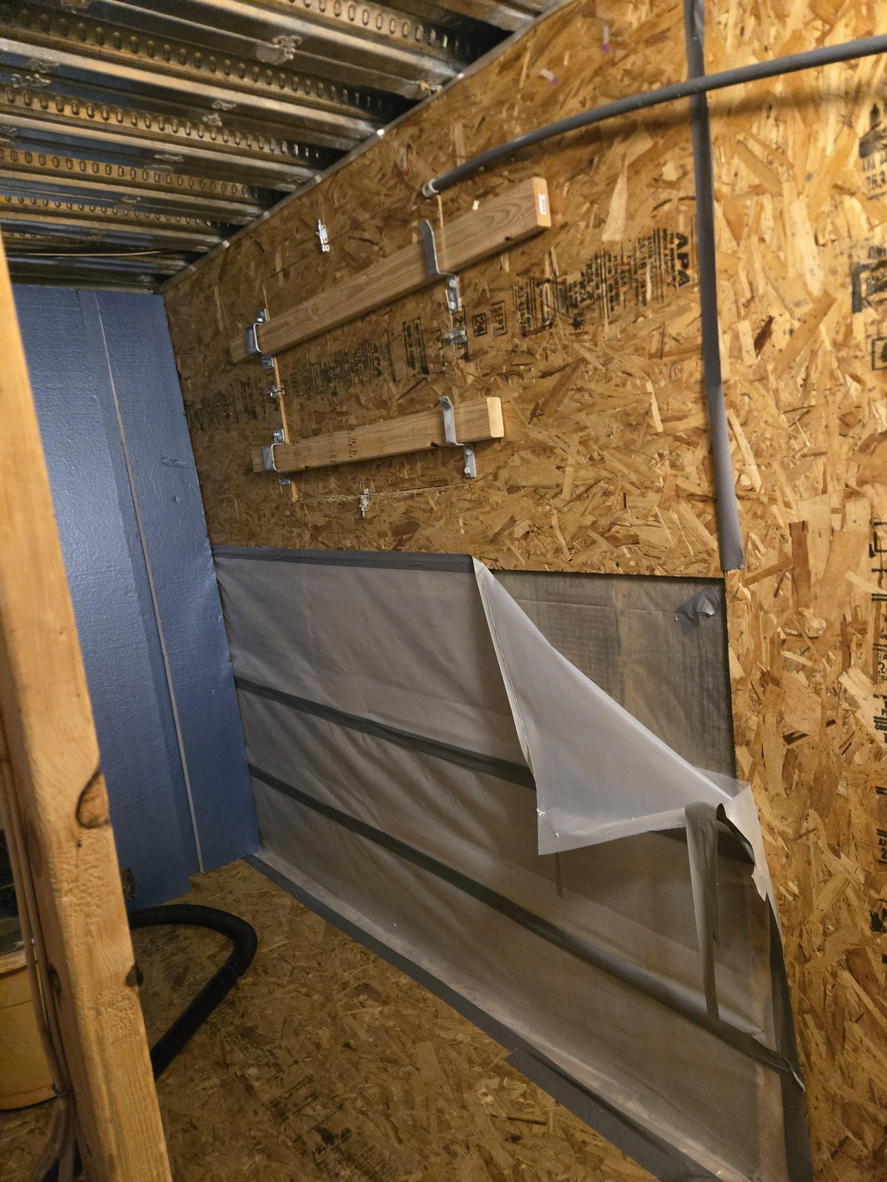 Interior of a firewood storage area showing exposed OSB walls, blue insulation, and protective plastic sheeting, indicating ongoing water damage remediation and structural work.