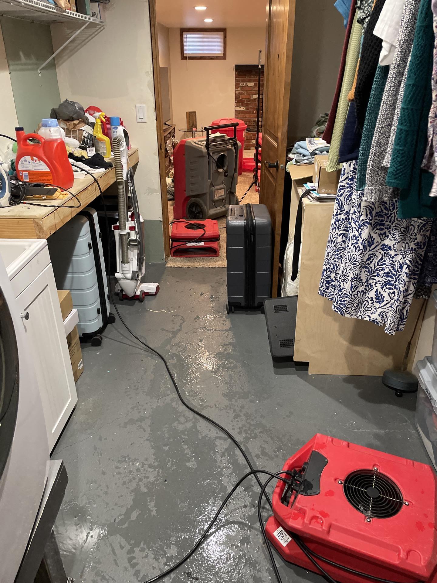 A basement utility room with standing water on the concrete floor, showing active water damage restoration with red air movers and a grey commercial dehumidifier.
