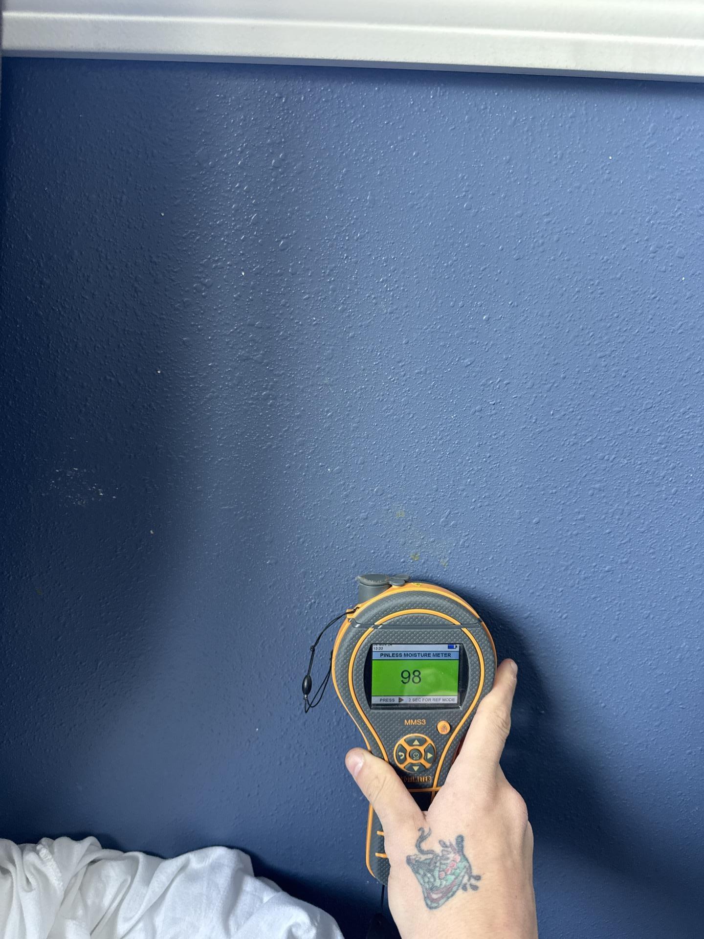Hand holding a moisture meter showing a reading of 98 on a blue wall, confirming persistent moisture in a different area of the damaged bedroom.