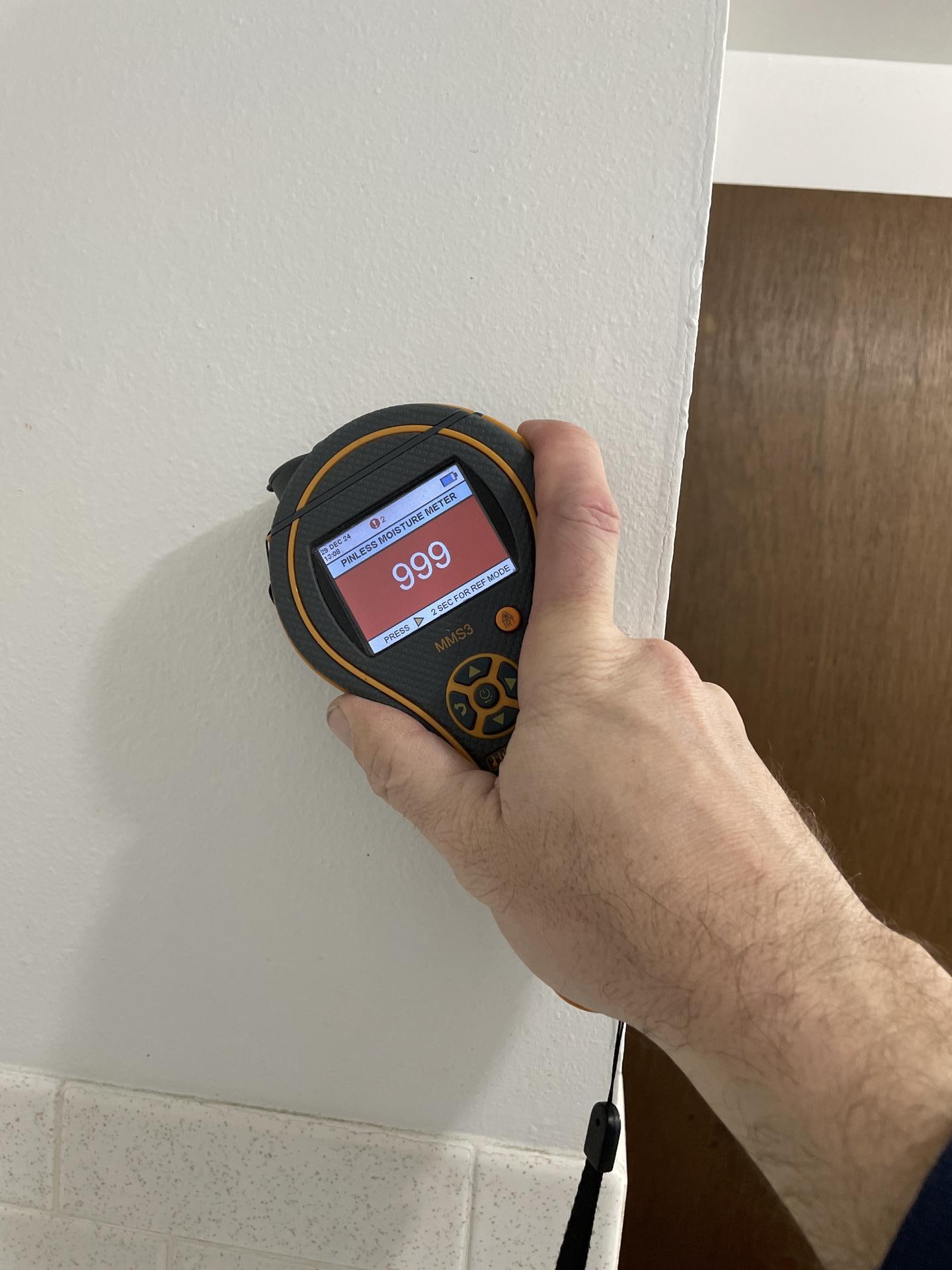 A restoration technician uses a moisture meter showing '999' on a main floor bathroom wall, indicating severe hidden water damage and saturation.