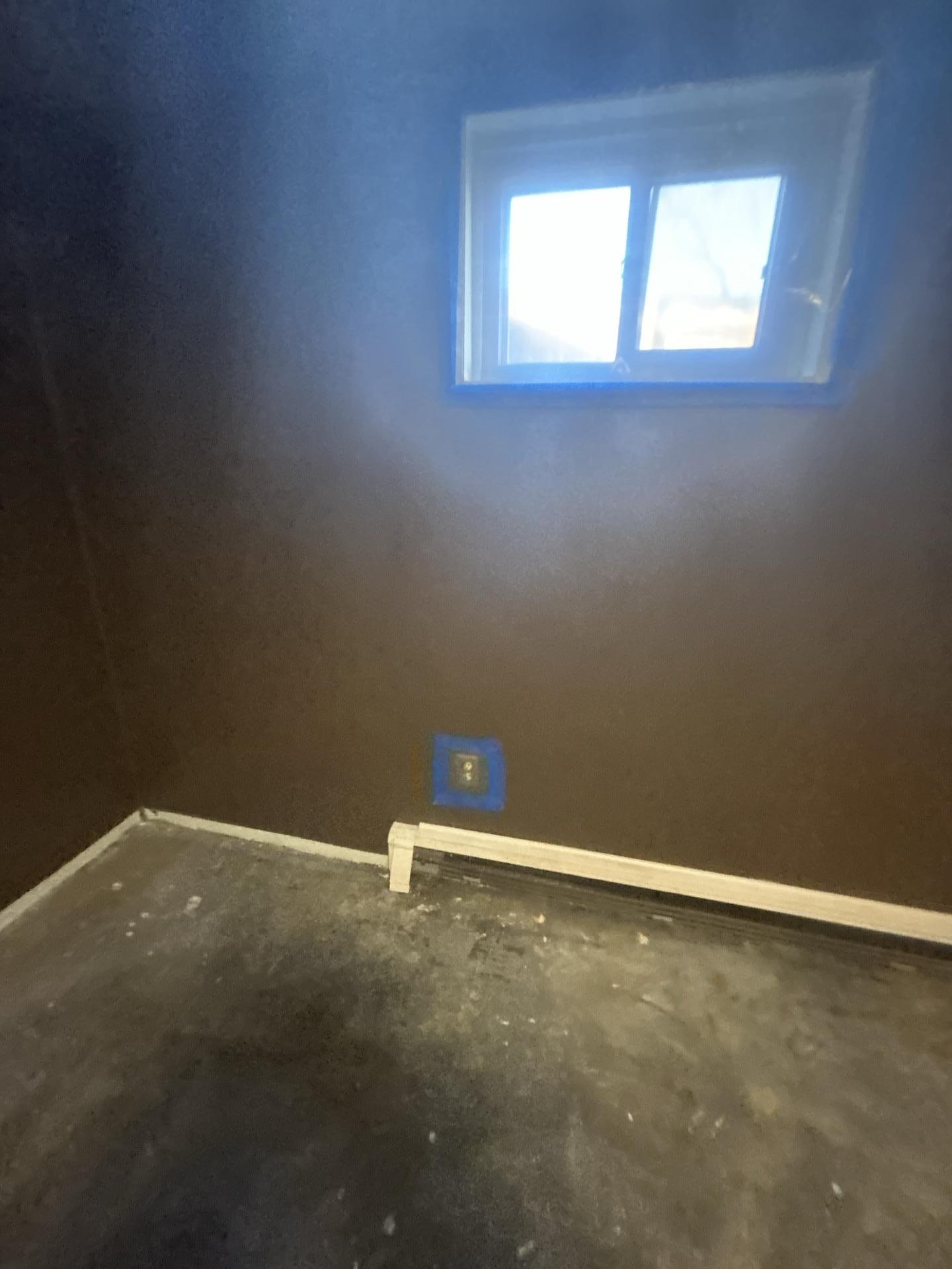 A close-up of a basement corner showing a window with blue protective tape, an electrical outlet, and a baseboard heater, post-water damage restoration.