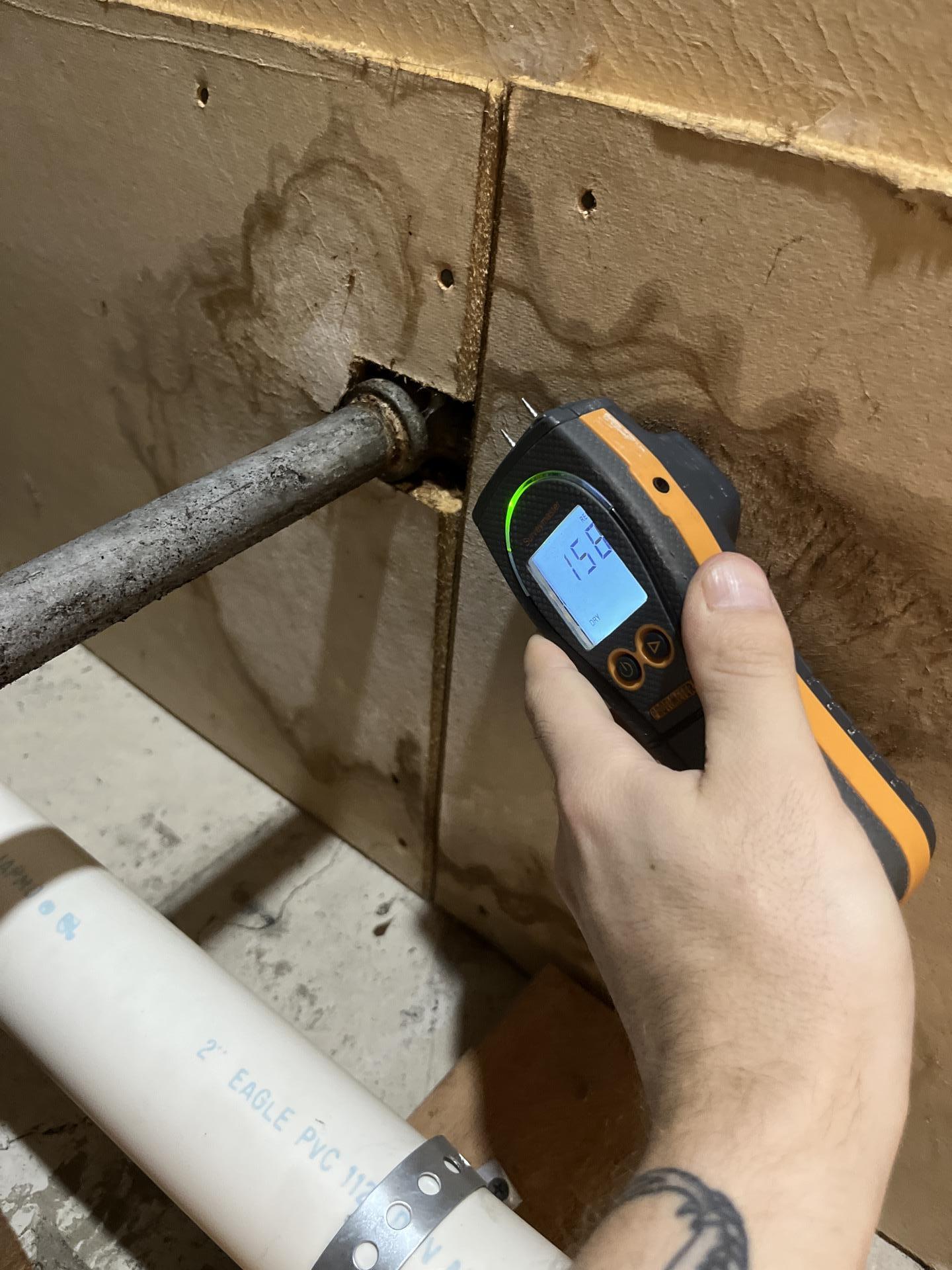 Moisture meter showing a high reading (15.8) on water-stained basement paneling with a pipe, indicating past water intrusion.